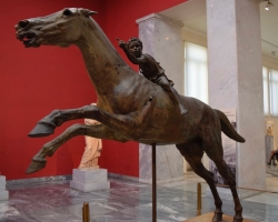 Artemision-jockeyn. Den väldiga hästen nästan flyger fram och den lilla pojken lutar sig framåt och manar på hästen för att öka farten ännu mer. Smidd helt i brons under hellenistisk tid, cirka 140 f Kr. Hittad i ett skeppsvrak norr om ön Euboea.